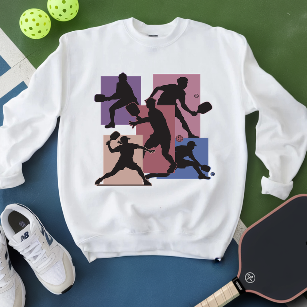 White sweatshirt with pickleball player design on a green and blue surface with pickleballs and a paddle.