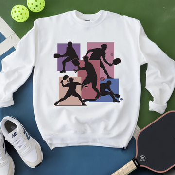 White sweatshirt with pickleball player design on a green and blue surface with pickleballs and a paddle.
